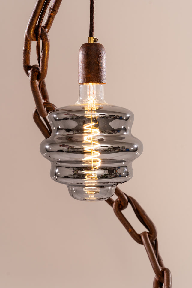 Dettaglio ravvicinato della lampadina vetro fumé con filamento acceso e cavo elettrico visibile tra le maglie della catena in ferro