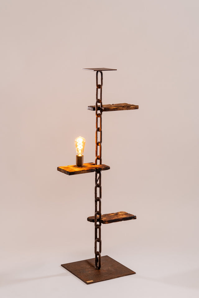 Dettaglio ravvicinato della lampadina accesa su ripiano in legno con superficie bruciata e catena metallica come colonna portante