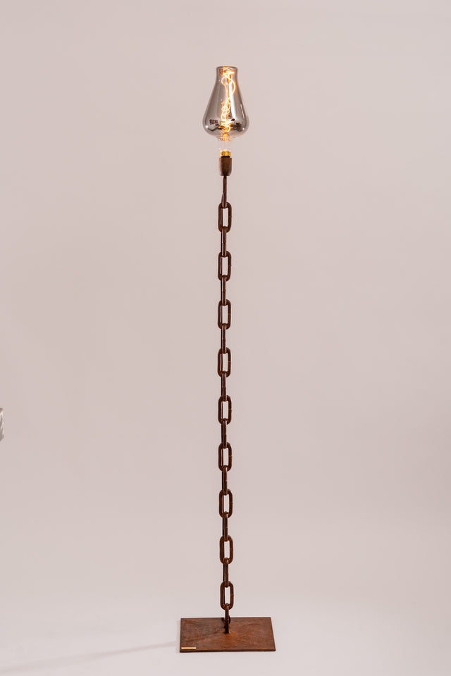 Lampada da terra Castena modello Hera, realizzata con catena navale verticale e base quadrata in metallo ruggine, con lampadina decorativa a filamento e vetro fumé