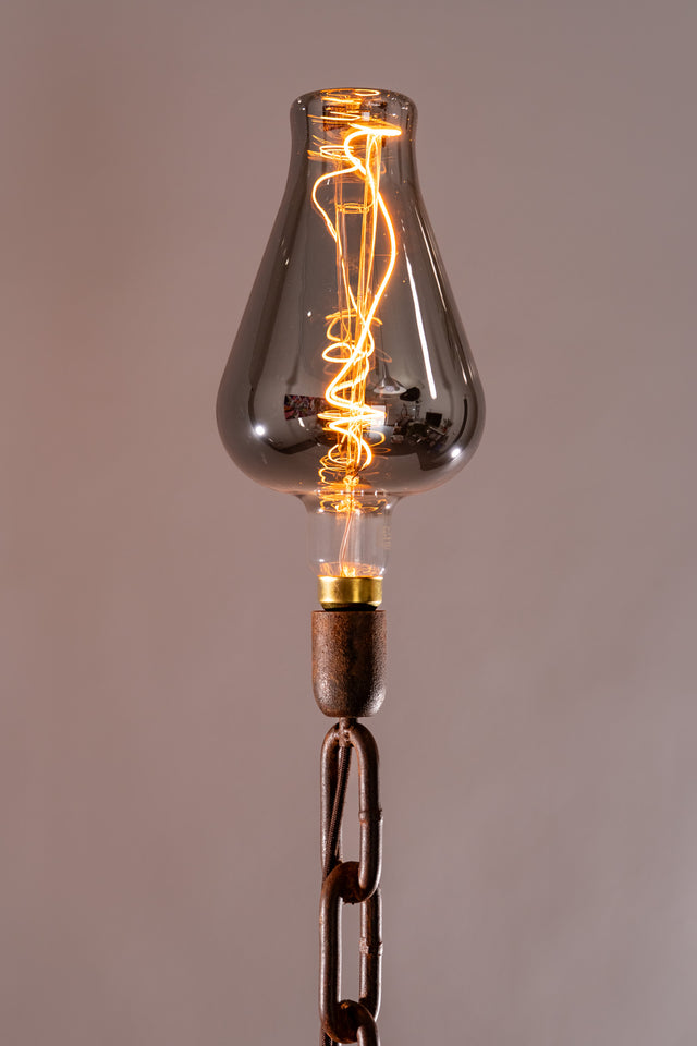 Dettaglio ravvicinato della lampadina fumé con filamento a spirale accesa, supportata da un portalampada ossidato e catena in ferro brunito