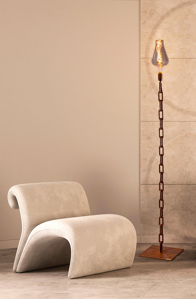 Ambientazione moderna con la lampada Hera accesa accanto a una poltrona chiara in velluto, su pavimento in cemento, stile industriale raffinato