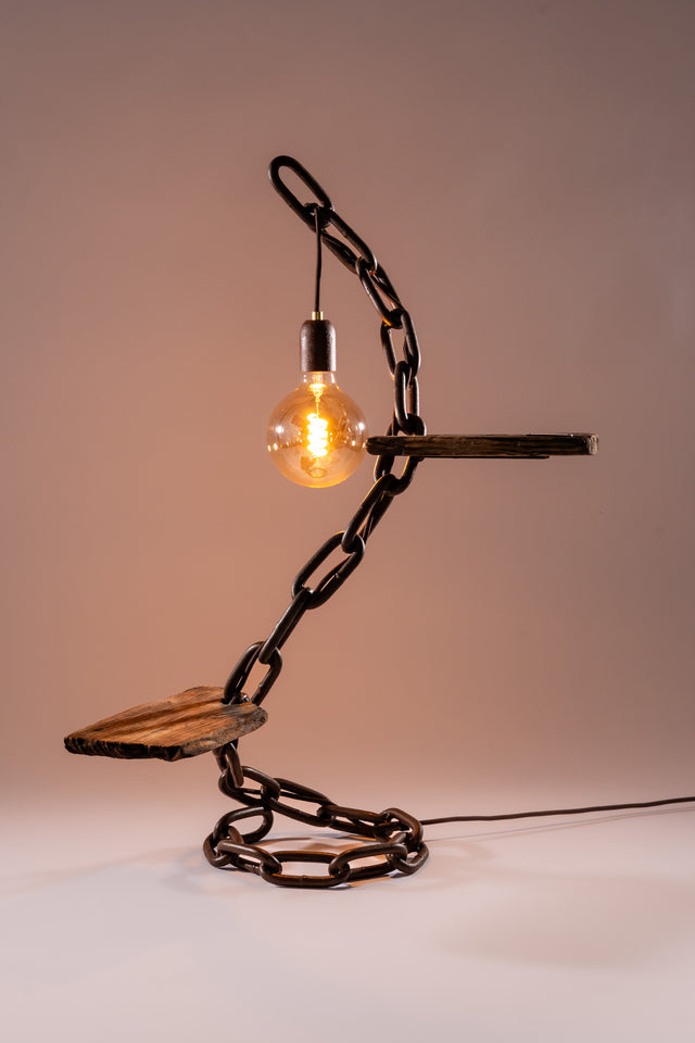 Lampada da tavolo con struttura scultorea in catena e legno, luce tonda calda e cavo elettrico visibile sul fondo, design Castena