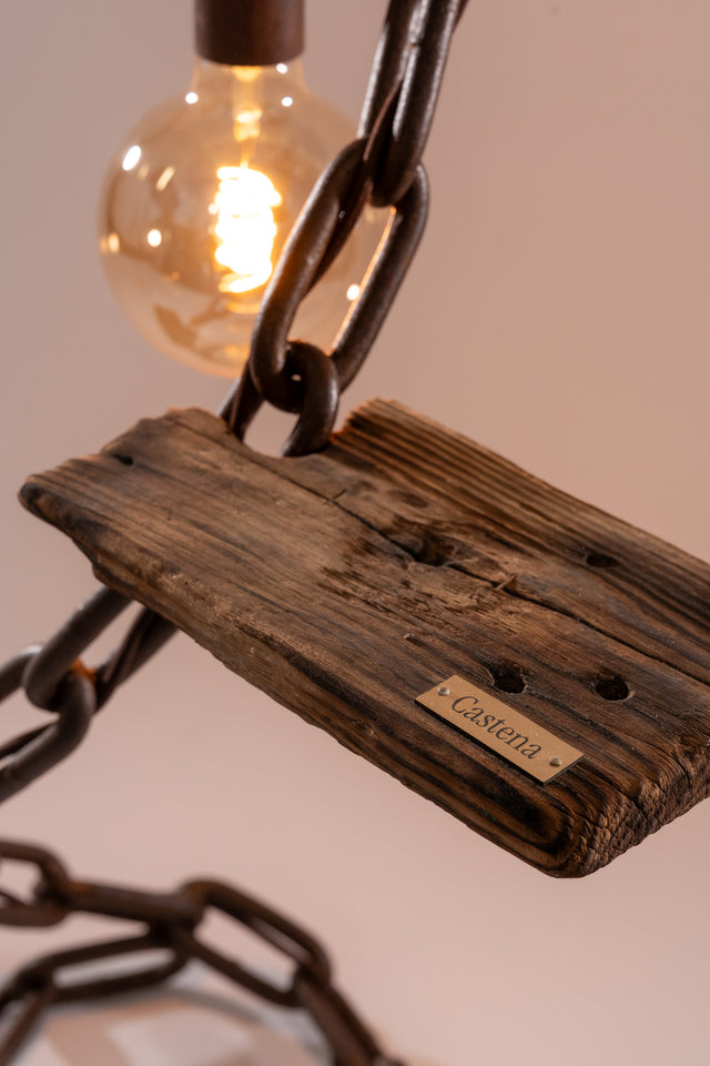 Dettaglio della placca in legno vissuto con logo Castena inciso, lampadina accesa sullo sfondo e catena metallica in evidenza