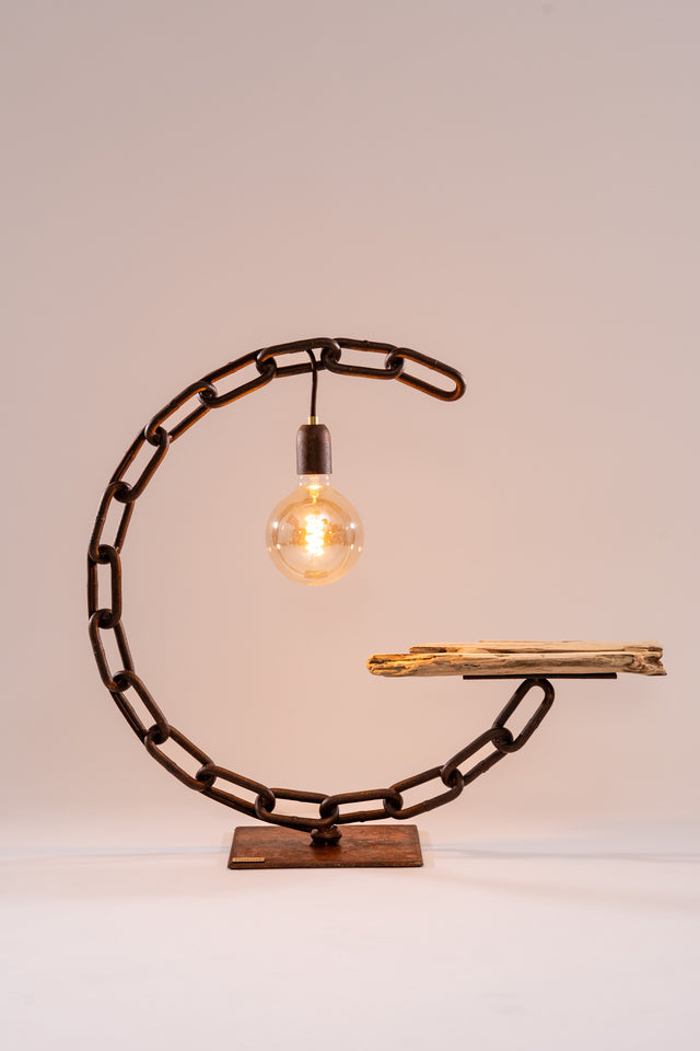 Lampada Polena con struttura a C in catena navale arrugginita, luce LED tonda e ripiano in legno grezzo, con cavo elettrico visibile alla base