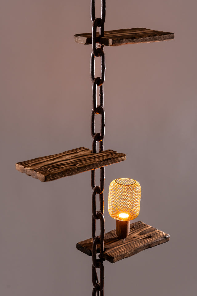 Dettaglio ravvicinato dei ripiani in legno vissuto e lampadina con copertura in rete metallica, sorretti da una catena verticale in ferro brunito